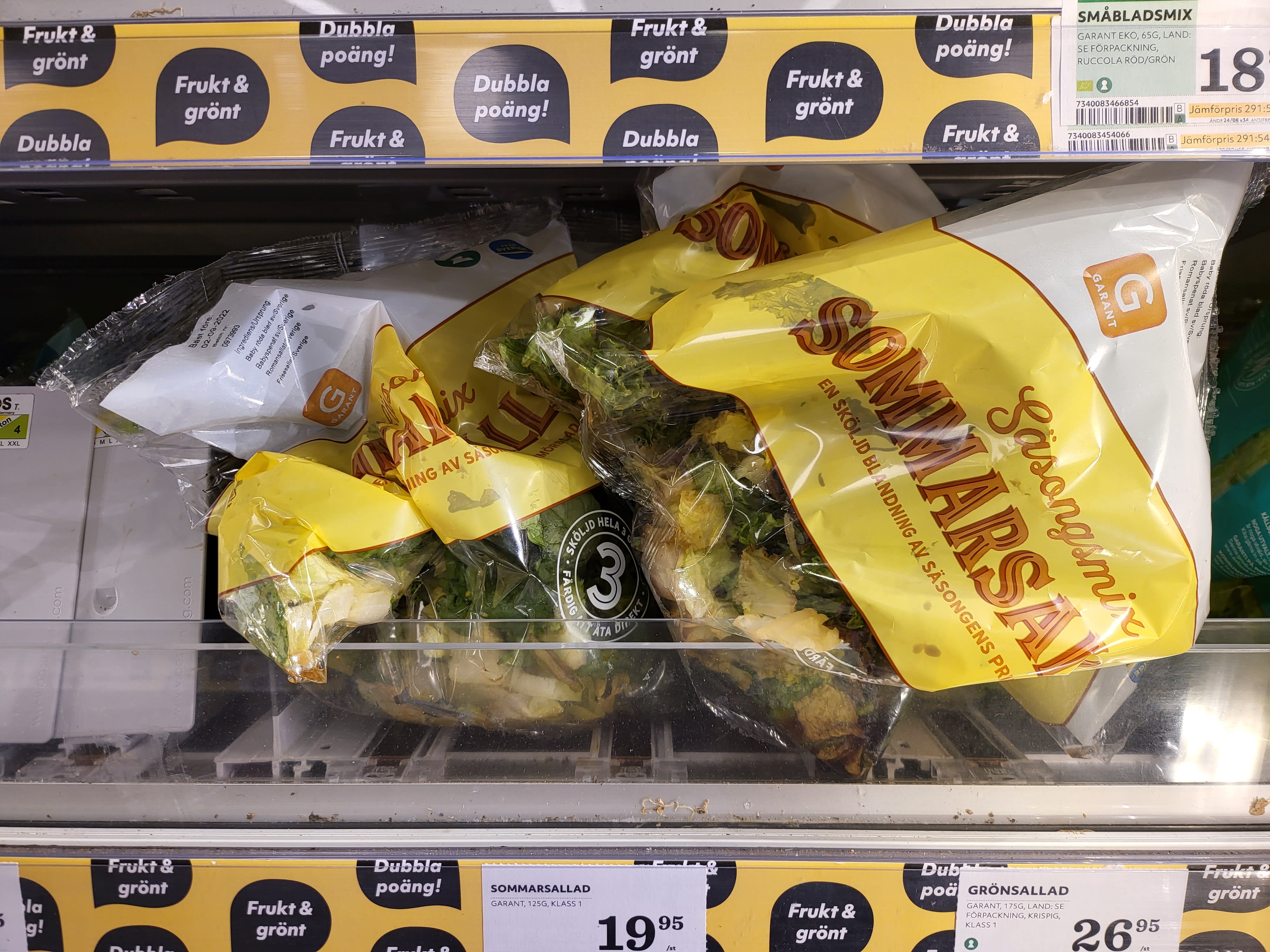 Påsar med sommarsallad ligger i en butikshylla med prislappar och gula skyltar om dubbla poäng.
