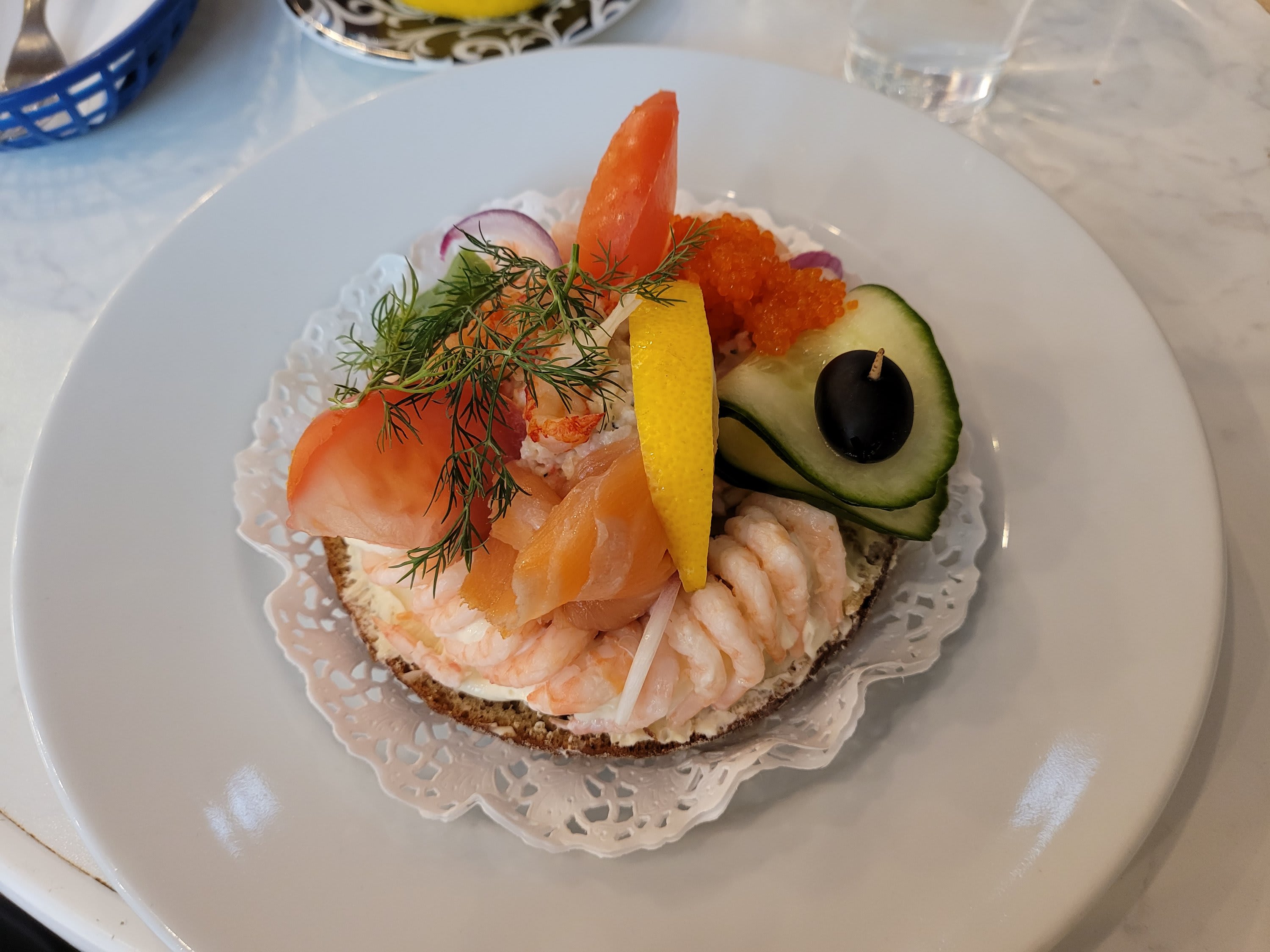Räkmacka med lax, rom, citron, dill, gurka och en svart oliv på en vit tallrik.
