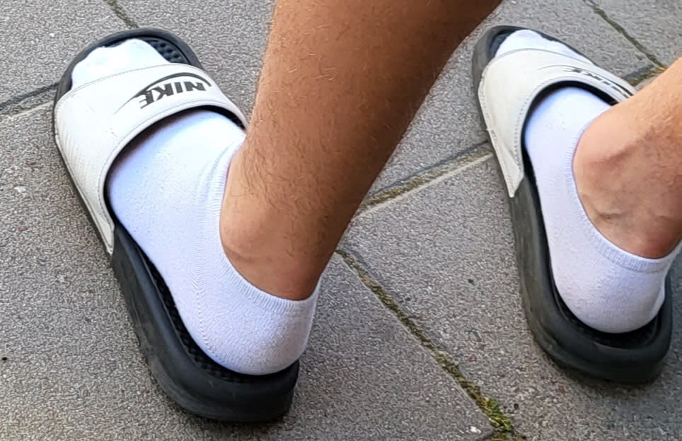 Två fötter i vita ankelstrumpor och svarta badtofflor med vita Nike-remmar på grå markplattor.