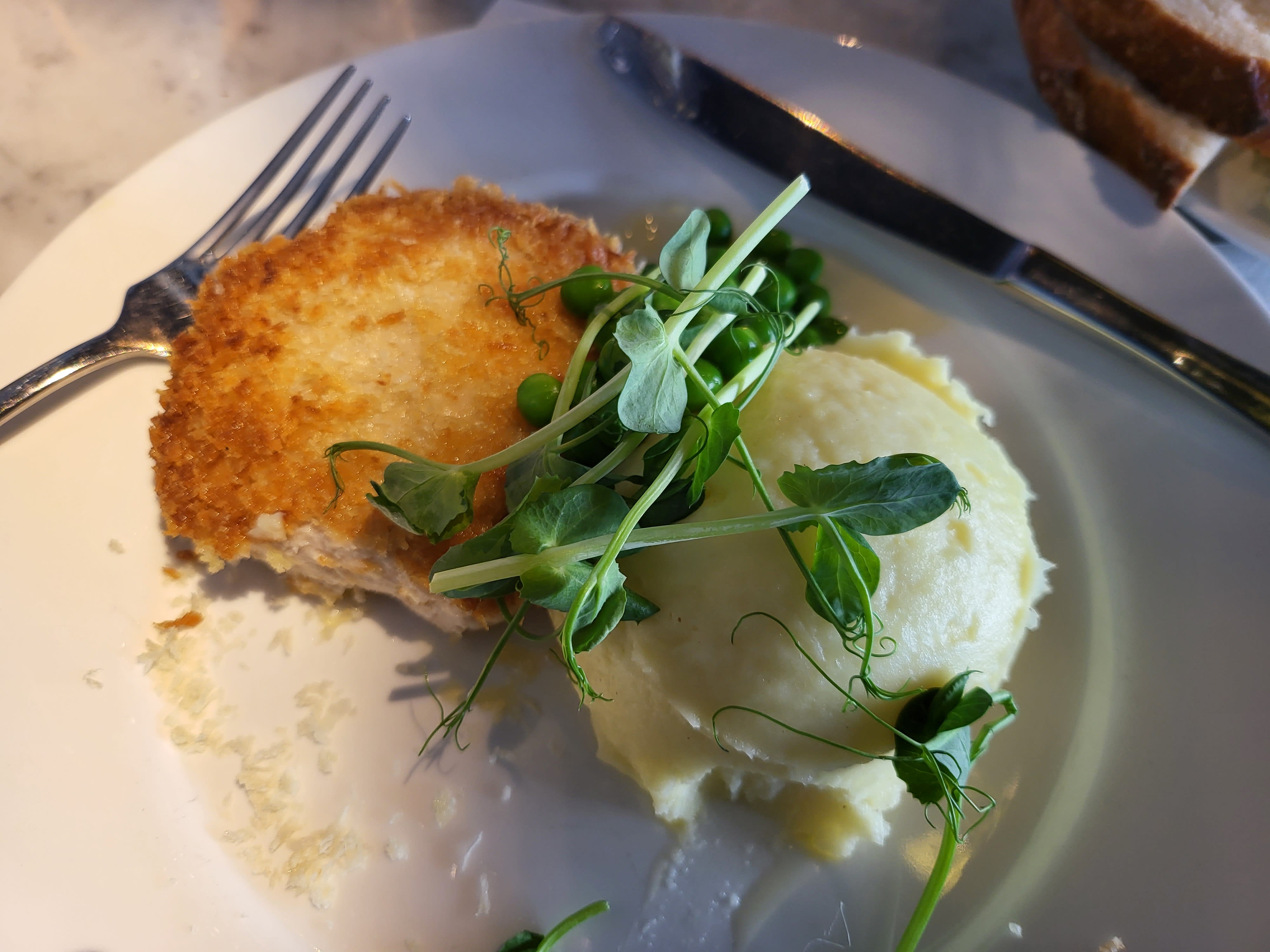 En tallrik med en gyllenbrun panerad biff, potatismos, gröna ärtor och ärtskott, med bestick bredvid.