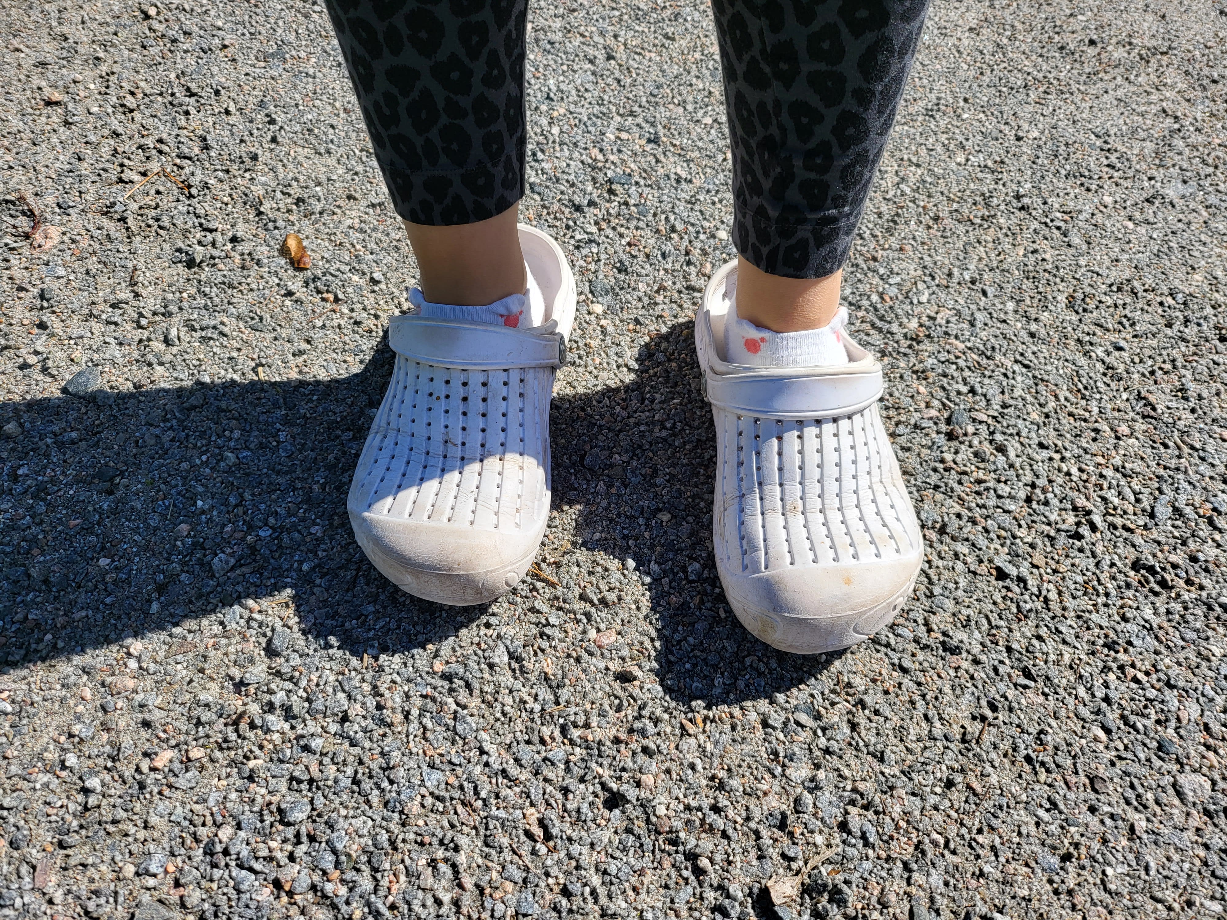 Fötter i vita perforerade tofflor och strumpor står på grus, under mörka leopardmönstrade leggings.