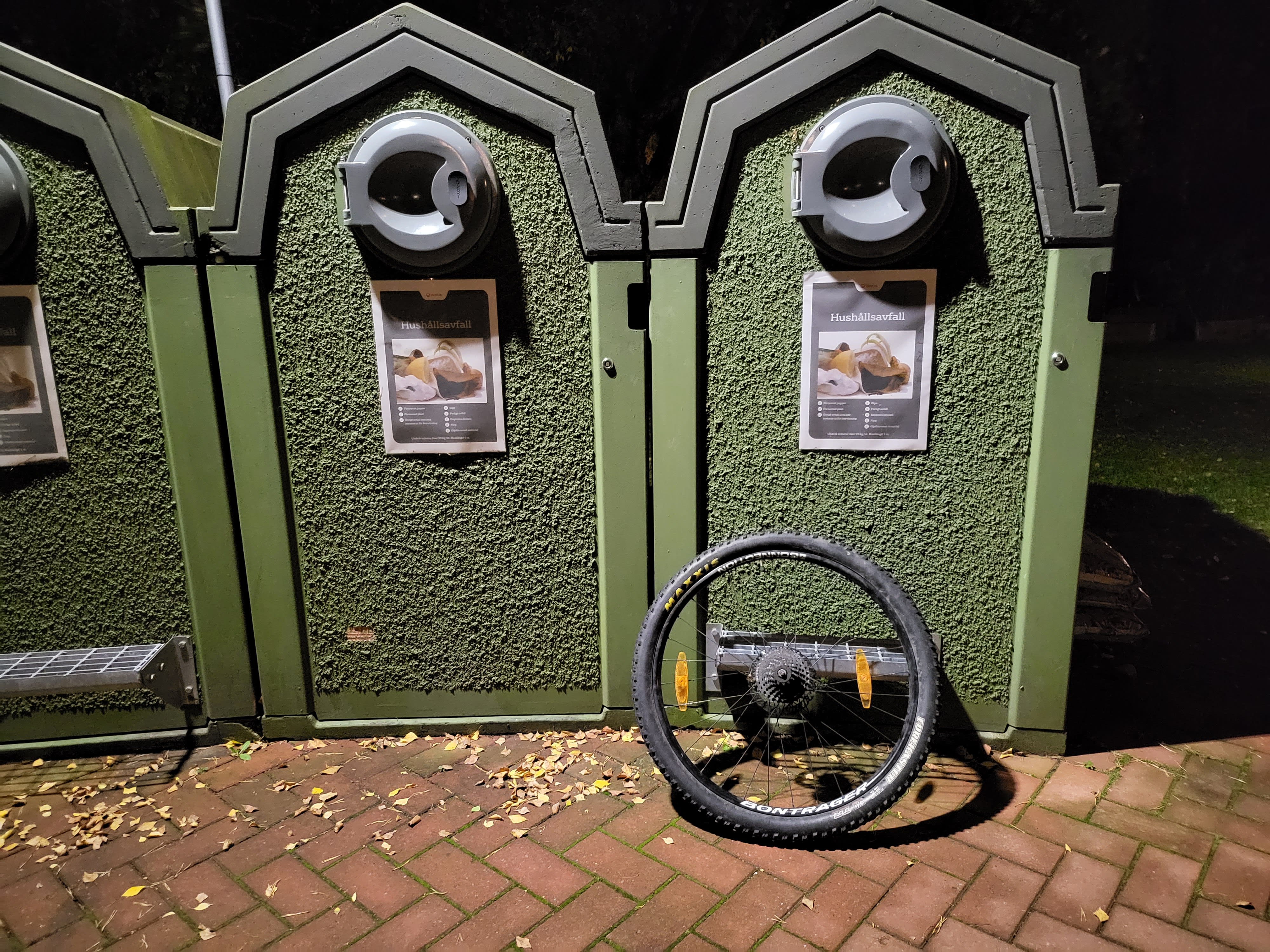 Ett löst cykelhjul lutar mot en grön sopbehållare för hushållsavfall på en tegelbelagd yta.