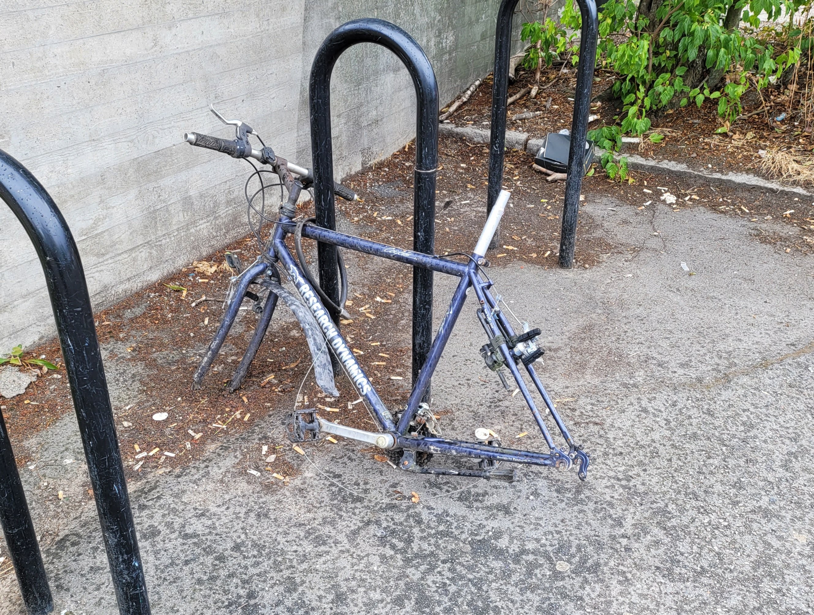 En cykelram utan hjul och sadel sitter fastlåst vid ett svart cykelställ intill en betongvägg.