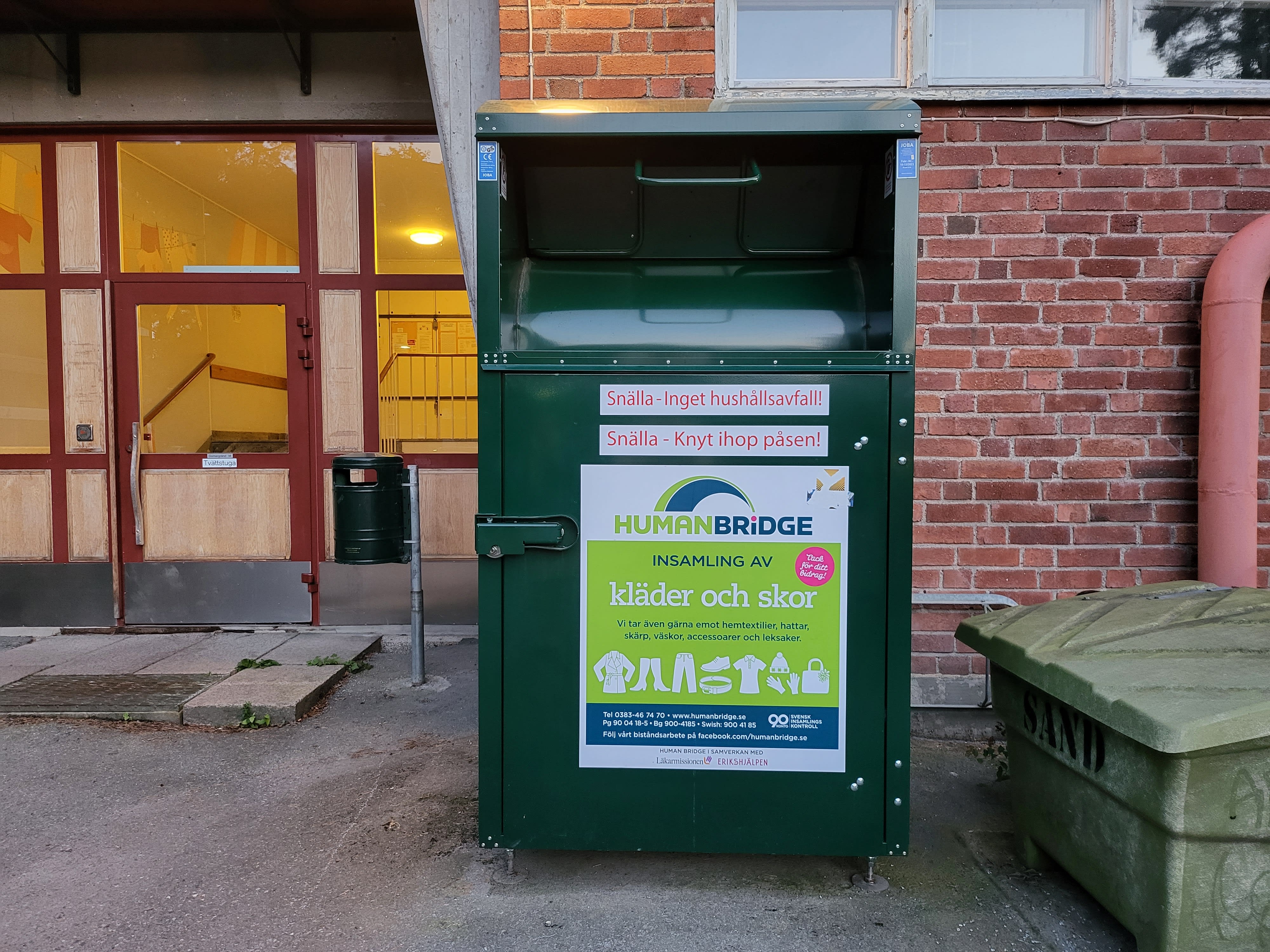 En klädinsamlingsbehållare med skyltarna ”Snälla - Inget hushållsavfall!” och ”Snälla - Knyt ihop påsen!” står utanför en tegelbyggnad.